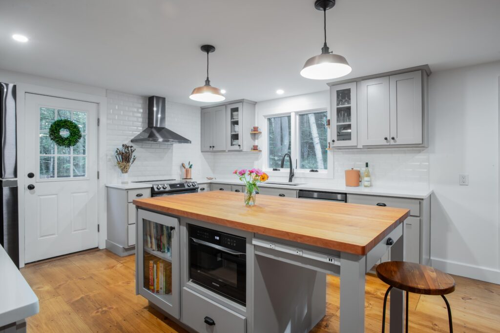 A gray and white kitchen with a center island with butcher block counter and two pendant lights. White subway tile backsplash and wood floors.