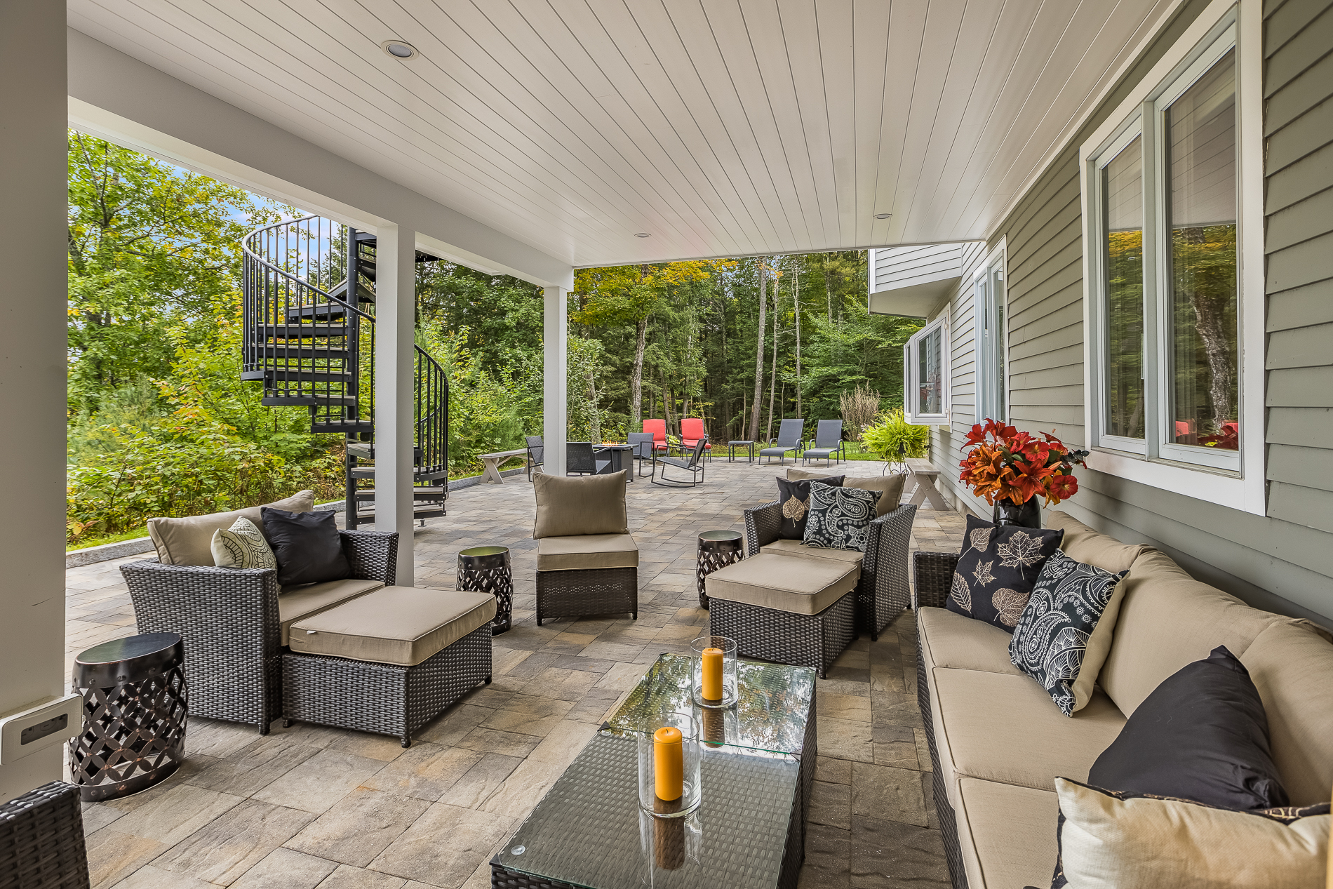 A remodeled covered patio with wicker furniture, forest in background, and black spiral metal staircase leading up to a balcony above.