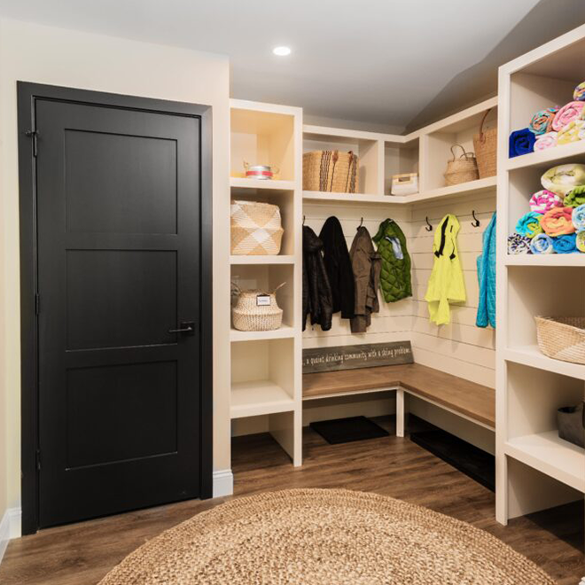 Neatly organized new mudroom with white shelving, woodgrain bench seat, wood floors, and black interior door.