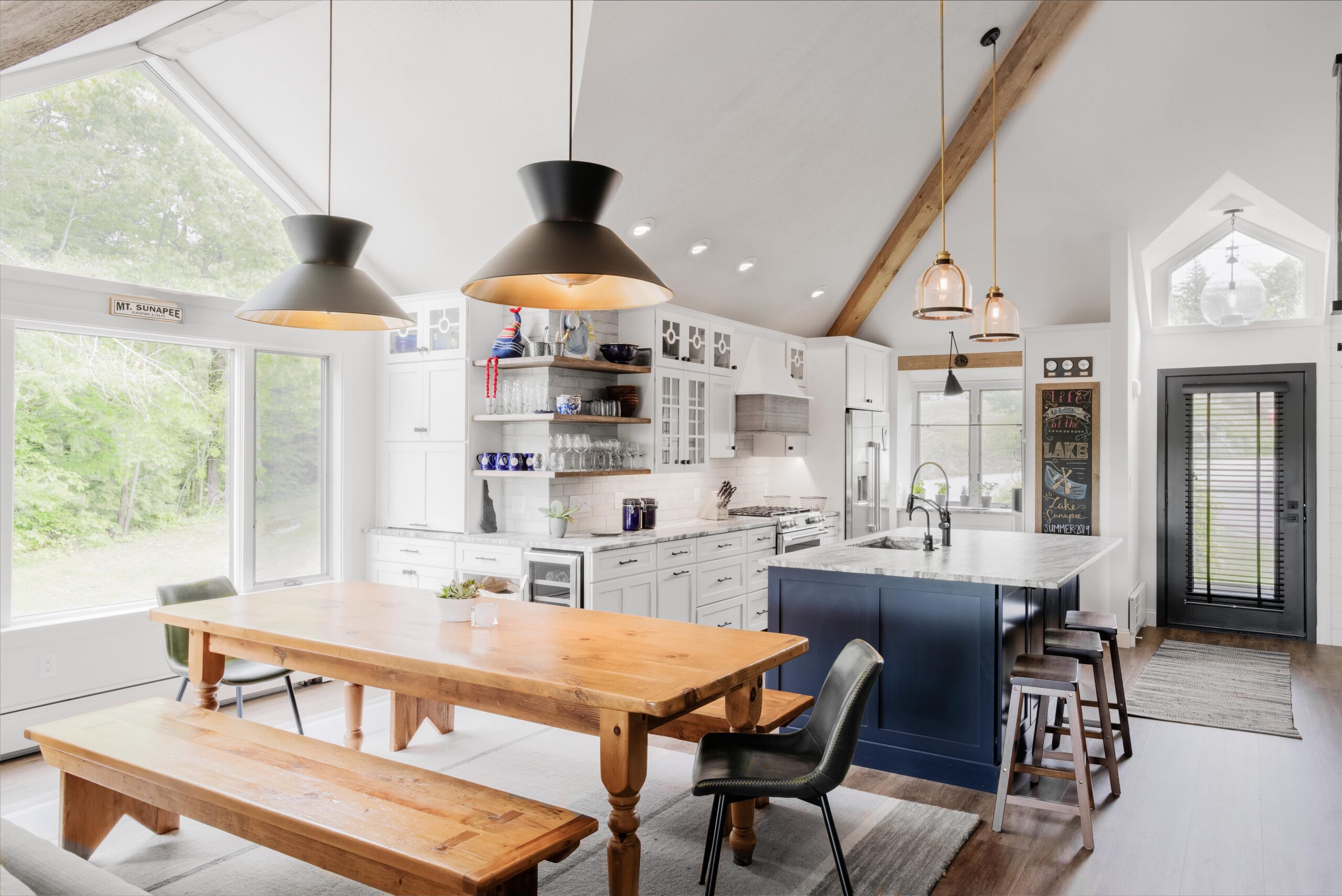 Whole-home remodel with open floor plan with white cabinets and large wood picnic-style dining table next to large picture windows. Light wood floors and vaulted white ceiling.