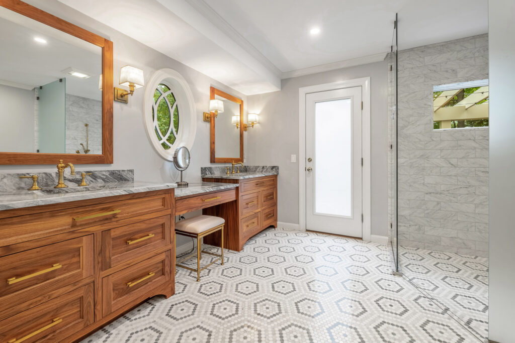 Remodeled bathroom design with an accessible walk-in shower, woodgrain dual vanity with gray marble counters, and small-patterned white and gray tile floor.