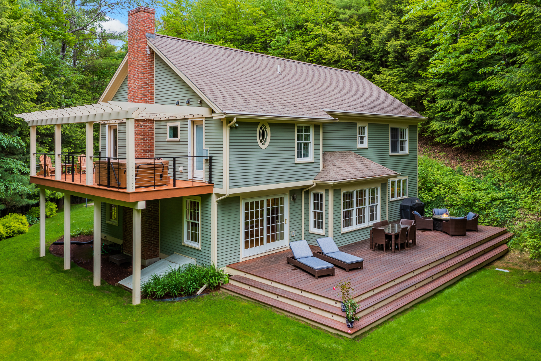 Exterior of a large two-story home with a finished basement that leads to a deck. Also has an upper deck on left side. Home has green siding and is surrounded by trees.