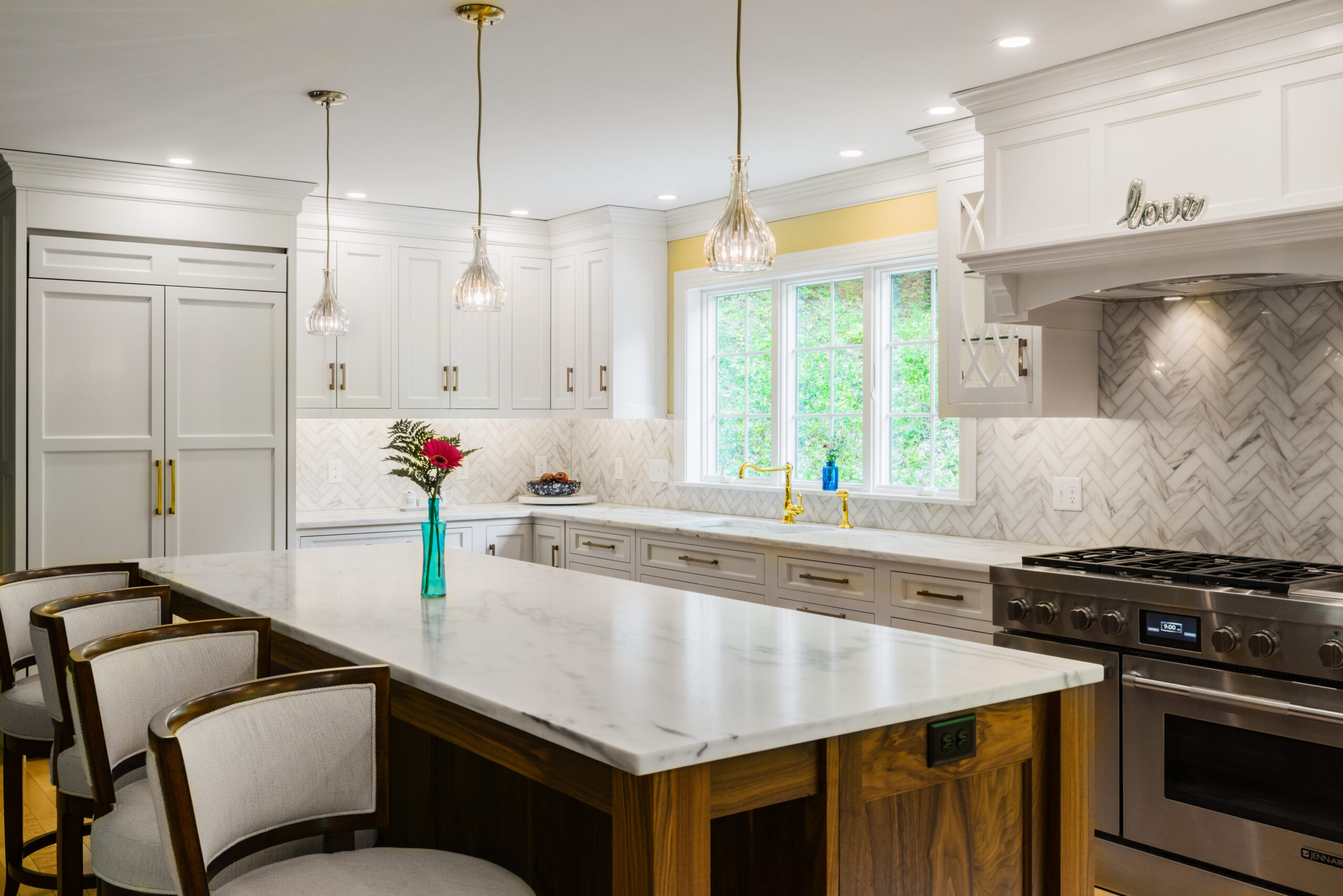 White marble countertop kitchen island with bar stool seating and teal vase with one pink flower. White cabinets along back wall, with windows over sink and white/gray herringbone backsplash.