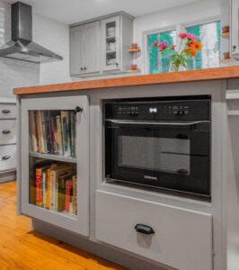 Custom gray cabinetry built into a kitchen island with butcher block counter top. Island includes built-in black microwave, glass-enclosed bookshelf, and a large drawer.