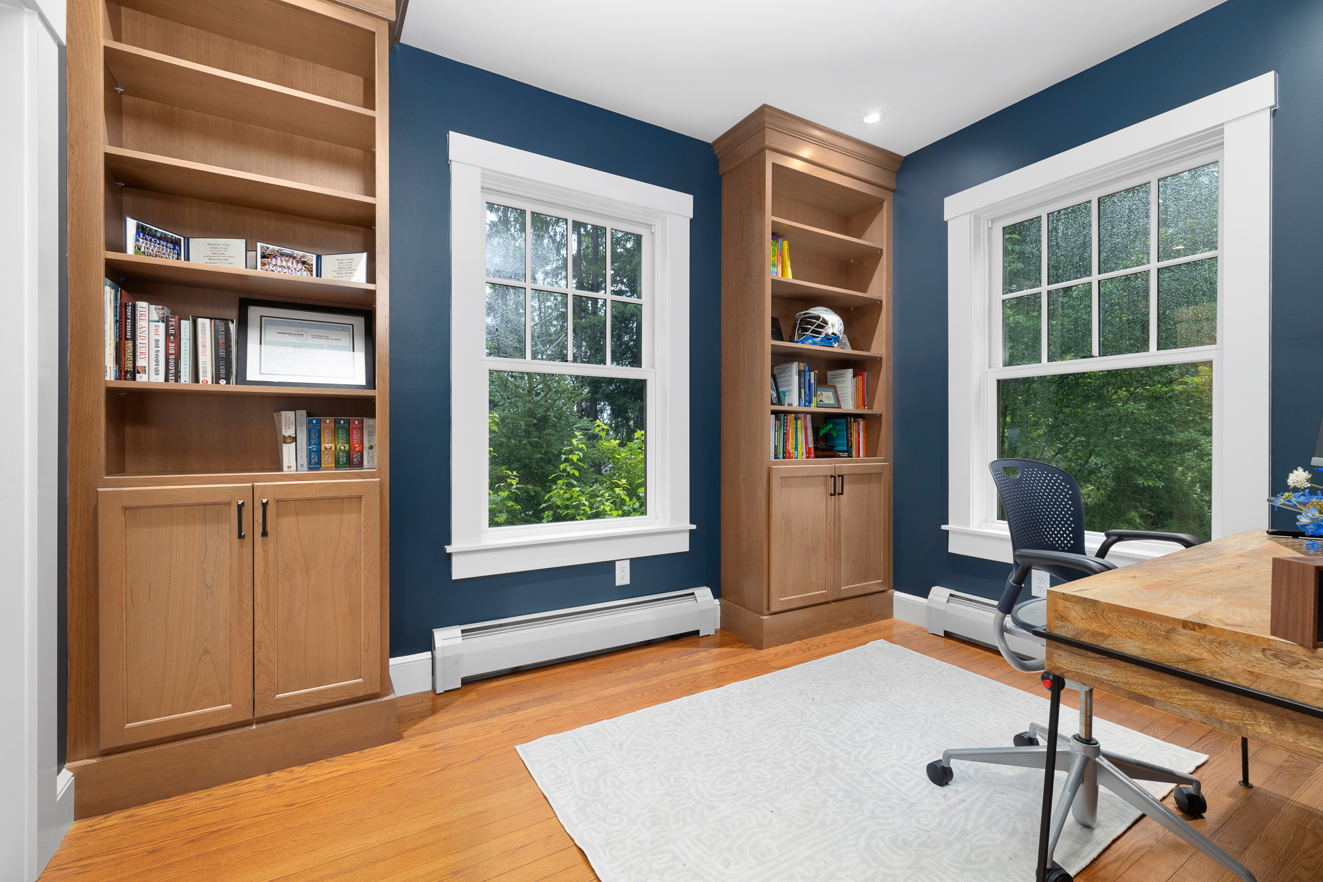 Remodeled home office with bookcases, wood floor, and two large double-hung windows with grids on top. Dark blue walls, and woodgrain desk in right corner.