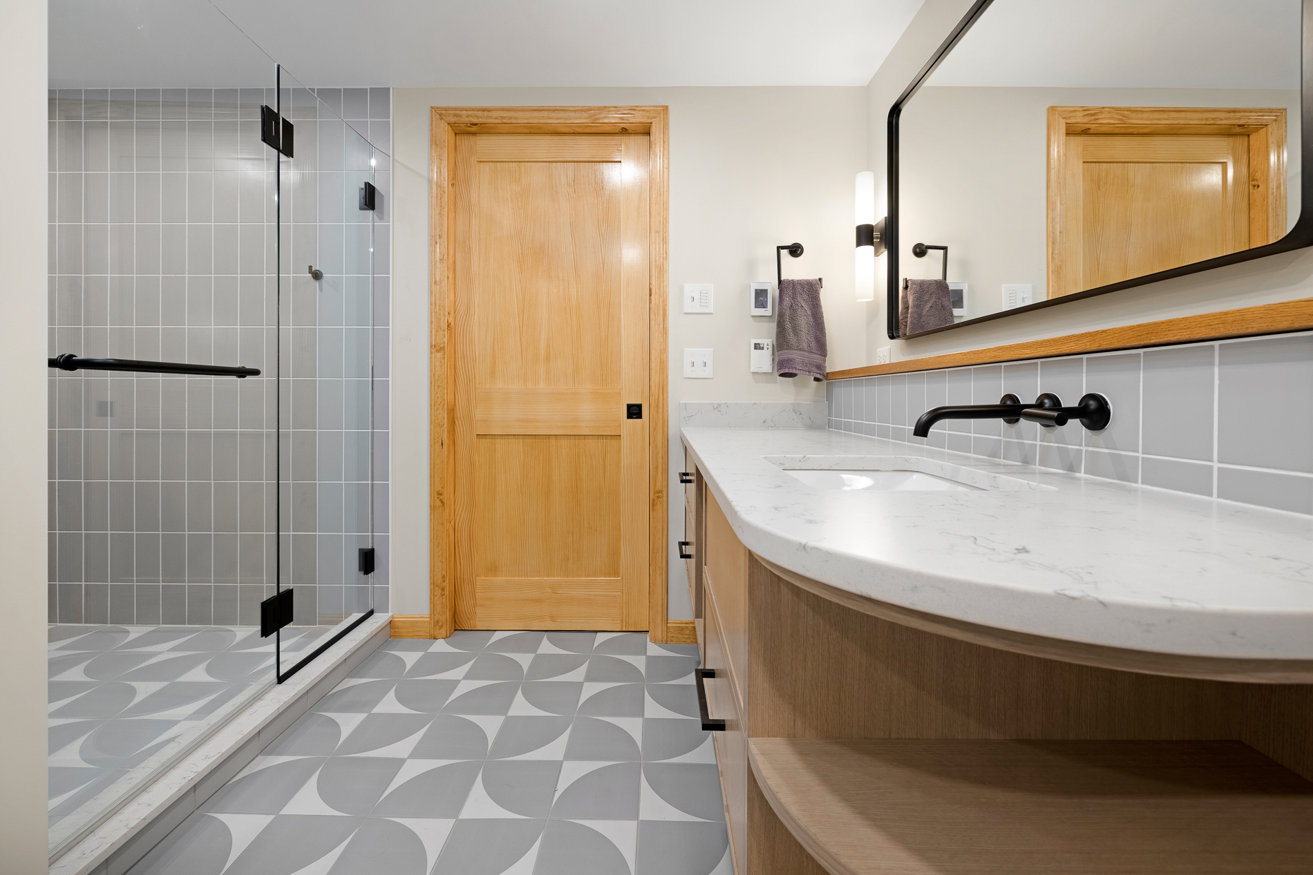 Modern bathroom with white granite counter, white and gray tile floor, glass-enclosed shower, and black fixtures.